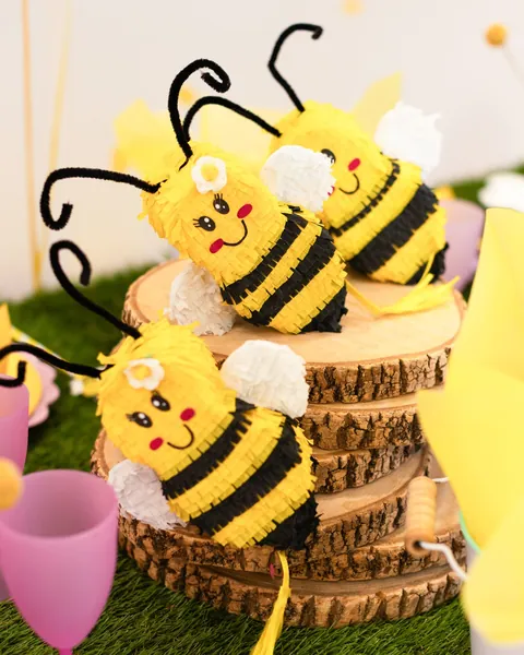 Tres piñatas de abeja pequeñas sobre rodajas de tronco, con rayas amarillas y negras, alas blancas y antenas rizadas.