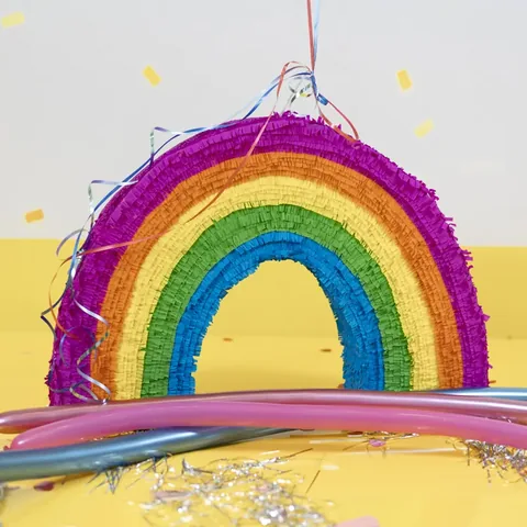 Piñata con forma de arcoíris, con flecos morados, naranjas, amarillos, verdes y azules, sobre una mesa amarilla con globos alargados y confeti.