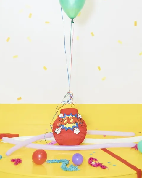 Piñata roja de superhéroe con un cartel «¡BOOM!», sobre el suelo de un estudio amarillo, bajo tres globos azul, rojo con lunares y verde.
