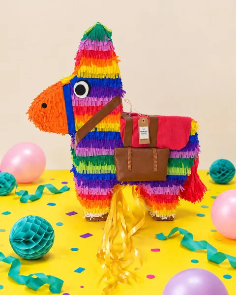 Piñata de burro con flecos de todos los colores del arcoíris, hocico naranja y alforjas marrones, sobre una mesa amarilla con globos, farolillos de papel turquesa, serpentinas y confeti.