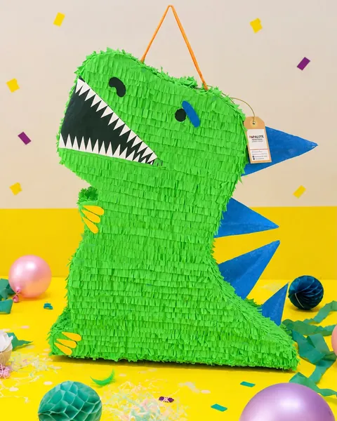 Piñata de dinosaurio verde de flecos, con la boca abierta y dientes blancos, uñas amarillas y púas azules en el lomo, sobre el suelo de un estudio amarillo con globos, bolas de papel y confeti.