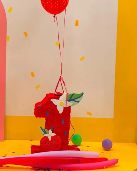 Piñata roja con forma del número 1, con una flor blanca y hojas verdes, en una fiesta con globos rojos, banderines y un fondo rosa y amarillo.